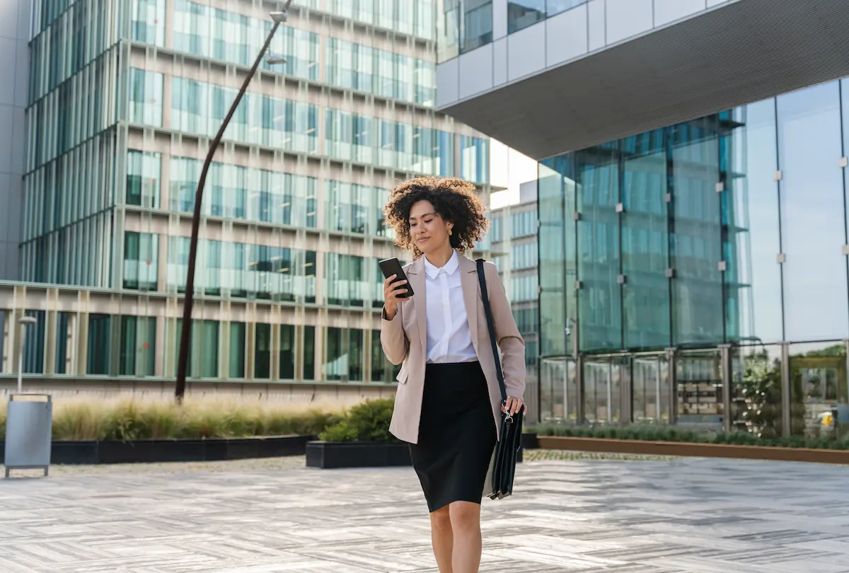 Businessfrau vor zwei Hochhäusern mit Aktentasche und Handy.